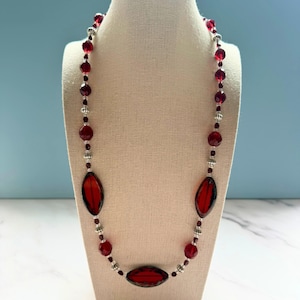 May include: A red beaded necklace with oval and round beads. The necklace features a mix of dark red and silver-colored beads, with three large oval-shaped red beads as a focal point. The necklace is displayed on a mannequin.