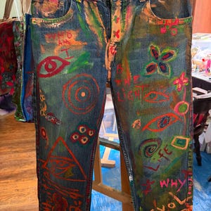 May include: Blue denim jeans featuring a Levi's brand label at the waist and hand-painted designs in vibrant colors. The artwork includes various symbols, eyes, geometric shapes, and handwritten text such as "WHO AM I", "BE THE LIGHT", and "LIFE".
