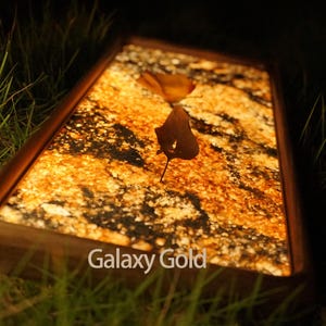 Op de afbeelding: Een rechthoekig houten frame met een verlicht, goud en zwart marmer oppervlak. Het oppervlak wordt van achteren verlicht, waardoor een warme gloed ontstaat. De tekst "Galaxy Gold" is zichtbaar aan de onderkant van het frame.