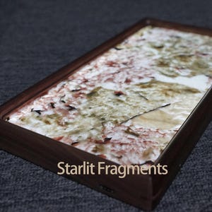 May include: A rectangular tray with a dark wood frame and a white, pink, and brown marbled surface. The tray has the text "Starlit Fragments" printed on the bottom edge.