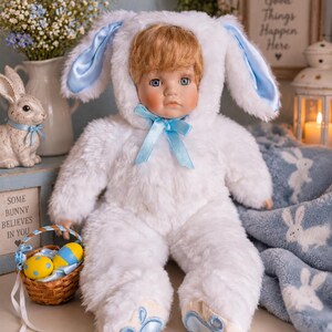 May include: A porcelain doll with light brown hair and blue eyes wearing a white plush bunny costume with long ears, a blue satin ribbon bow at the neck, and blue paw pad accents on the feet. The doll has a soft, fuzzy texture and a serene expression.
