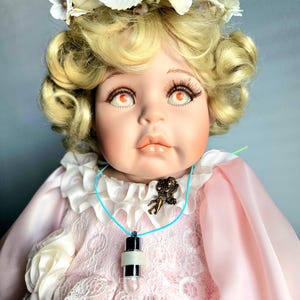 Könnte beinhalten: Eine Porzellanpuppe mit blonden Locken und einem Blumenkranz. Die Puppe hat große Augen, lange Wimpern und trägt ein rosa Kleid mit Spitzendetails. Eine Halskette mit Anhänger und einem Glühbirnen-Charm hängt um den Hals der Puppe.