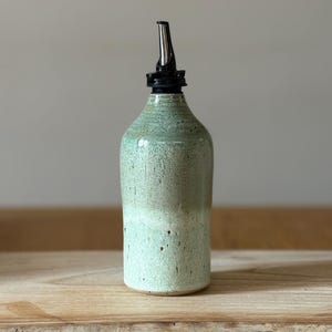 May include: A handmade ceramic bottle with a light green glaze and a black spout. The bottle has a rounded shape and a textured surface. The spout is made of metal. The bottle is on a wooden surface. The bottle is approximately 20 cm tall.