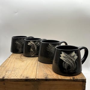 May include: Five black ceramic mugs with white abstract designs. The mugs are arranged in a row on a wooden surface.