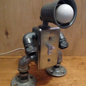 May include: A unique, industrial-style lamp crafted from metal pipes and fittings. The lamp features a light bulb in a spotlight housing, a metal switch plate, and a toggle switch. The lamp is mounted on a wooden surface.