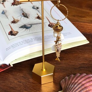 Könnte beinhalten: Goldene Muschel-Ohrringe mit einem goldenen Kreis und einem baumelnden Muschel-Anhänger. Der Anhänger ist mit einer Muschel, einer Perle und einem kleinen goldenen Fisch verziert. Die Ohrringe werden auf einem goldenen sechseckigen Ständer präsentiert.