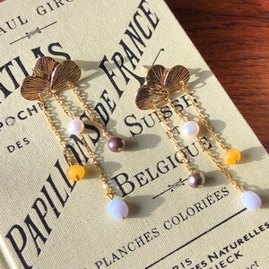 May include: Gold-coloured dangle earrings with a clover-shaped top and delicate chain. Each earring features a series of colourful beads in white, yellow, and purple. The earrings are displayed on a vintage book.