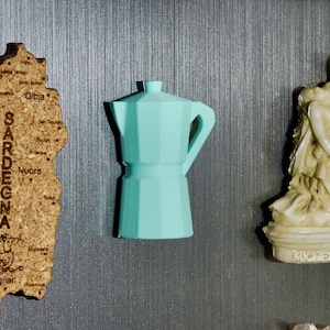 May include: A light blue ceramic coffee pot with a handle on the right side. The coffee pot is on a silver background with a cork map of Sardinia and a white ceramic sculpture of a woman.