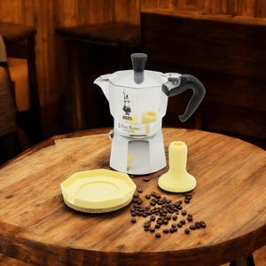Può includere: Una caffettiera Bialetti Moka Express argentata con manico nero e base in silicone gialla. La caffettiera è appoggiata su un tavolo di legno con un sottobicchiere in silicone giallo e chicchi di caffè sparsi intorno.