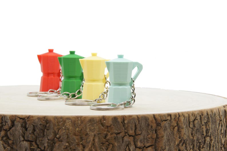 K&ouml;nnte beinhalten: Vier Miniatur-Espressokannen-Schl&uuml;sselanh&auml;nger in Rot, Gr&uuml;n, Gelb und Hellblau. Die Schl&uuml;sselanh&auml;nger liegen auf einer Holzoberfl&auml;che.