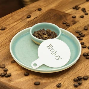 May include: A light blue ceramic dish with a smaller light blue bowl inside. The bowl is filled with coffee beans. A white stencil with the word "Enjoy" is on the dish. Coffee beans are scattered on a wooden surface.