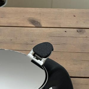 Op de afbeelding: Een glanzend roestvrijstalen deksel met een zwarte, getextureerde handgreep. Het deksel bedekt gedeeltelijk een zwart, getextureerd oppervlak. De afbeelding is geplaatst tegen een houten achtergrond.