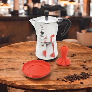 Könnte beinhalten: Eine silberne Bialetti Moka Express Espressomaschine zum Kochen auf dem Herd mit einem roten Silikonboden und einem roten Silikondrücker. Die Espressomaschine hat einen schwarzen Griff und einen schwarzen Knopf. Der Tamper steht auf einem Holztisch mit verstreuten Kaffeebohnen.