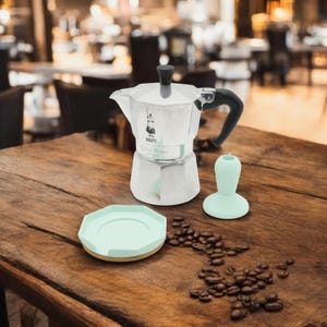 Op de afbeelding: Een zilveren Bialetti Moka Express koffiezetapparaat met een zwarte handgreep, een lichtgroene siliconen bekerhouder, een lichtgroene siliconen onderzetter en koffiebonen die verspreid liggen op een houten tafel.
