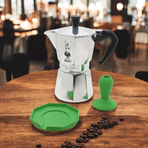 May include: Silver Bialetti Moka Express coffee maker with a black handle and green silicone base and drip tray. The coffee maker is on a wooden table with coffee beans scattered around it.