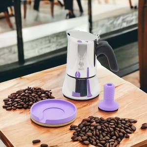 Op de afbeelding: Een espressomachine van roestvrij staal met een paarse siliconen basis en een paarse siliconen handvat. De espressomachine staat op een houten oppervlak met koffiebonen eromheen.
