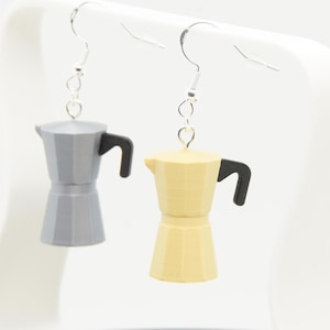 Può includere: Un paio di orecchini a forma di caffettiere in miniatura. Uno è grigio con un manico nero, l'altro è giallo con un manico nero. Entrambi hanno ganci per le orecchie color argento.