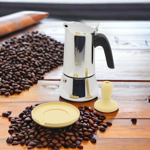 Op de afbeelding: Een zilveren espressomachine voor op het fornuis met een zwarte handgreep en een gele kunststof tamper. De espressomachine staat op een houten oppervlak met koffiebonen die eromheen verspreid liggen.