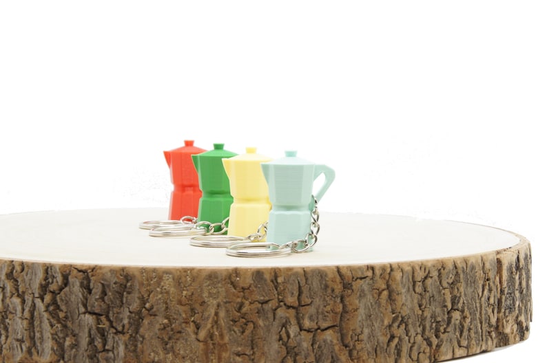 K&ouml;nnte beinhalten: Vier Miniatur-Kaffeekannen-Schl&uuml;sselanh&auml;nger in Rot, Gr&uuml;n, Gelb und Hellblau. Die Schl&uuml;sselanh&auml;nger sind auf einer Holzoberfl&auml;che abgelegt.