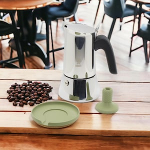Op de afbeelding: Een zilveren Bialetti Moka Express koffiezetapparaat met een groene siliconen basis en deksel. Het koffiezetapparaat staat op een houten oppervlak met een stapel koffiebonen ervoor.