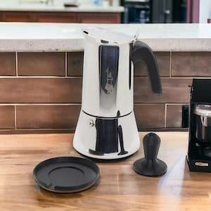 Op de afbeelding: Een zilveren espressomachine van roestvrij staal met een zwarte handgreep en een zwarte basis. De espressomachine staat op een houten oppervlak naast een zwarte tamper en een zwarte basis.