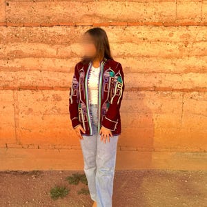 May include: A maroon velvet jacket with colourful embroidered geometric patterns. The jacket has a high collar and a button closure. The model is wearing light blue jeans and black heeled sandals.