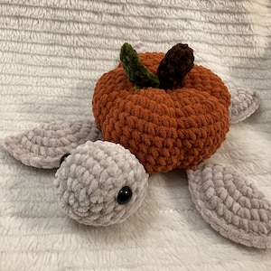 Puede incluir: Un juguete de tortuga de ganchillo con cabeza y aletas de color gris claro y un caparazón de calabaza naranja texturizado en la espalda. La calabaza está rematada con una pequeña hoja verde y un tallo marrón oscuro, y tiene ojos negros.
