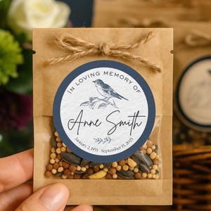 May include: A small, clear packet filled with seeds, sealed with a twine bow. A navy blue circular label reads "In Loving Memory of Anne Smith" with a bird illustration and dates. The packet is held in a hand.