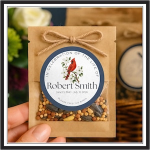 May include: A brown paper packet filled with birdseed, featuring a circular label with a red cardinal on a branch. The text reads: IN CELEBRATION OF THE LIFE OF Robert Smith JUNE 13, 1940 - JULY 31, 2026 PLEASE FEED THE BIRDS. A jute twine bow is tied at the top.