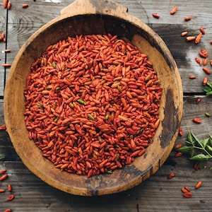 Op de afbeelding: Een houten kom gevuld met gedroogde rode chilipepers. De kom staat op een houten oppervlak met groene bladeren eromheen.