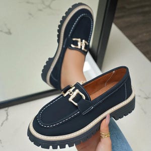 Puede incluir: Mocasines azul marino con hebilla dorada y costuras blancas. Los zapatos tienen una suela gruesa negra con ribete beige. La imagen muestra un par de zapatos, uno enfocado y el otro reflejado en un espejo.