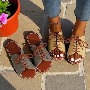 Op de afbeelding: Twee paar dames sandalen met een vetersluiting. Het ene paar is beige en het andere is groen. Beide paren hebben een bruine leren teenband en een witte zool.
