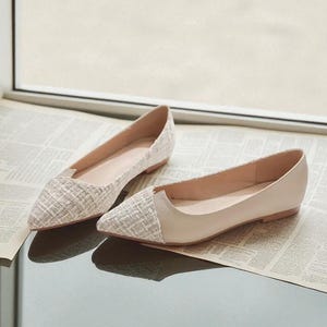 May include: A pair of beige and white flats with a pointed toe. The toe of each shoe has a textured, woven pattern, while the rest of the shoe is a smooth, solid beige. The shoes are displayed on a newspaper.