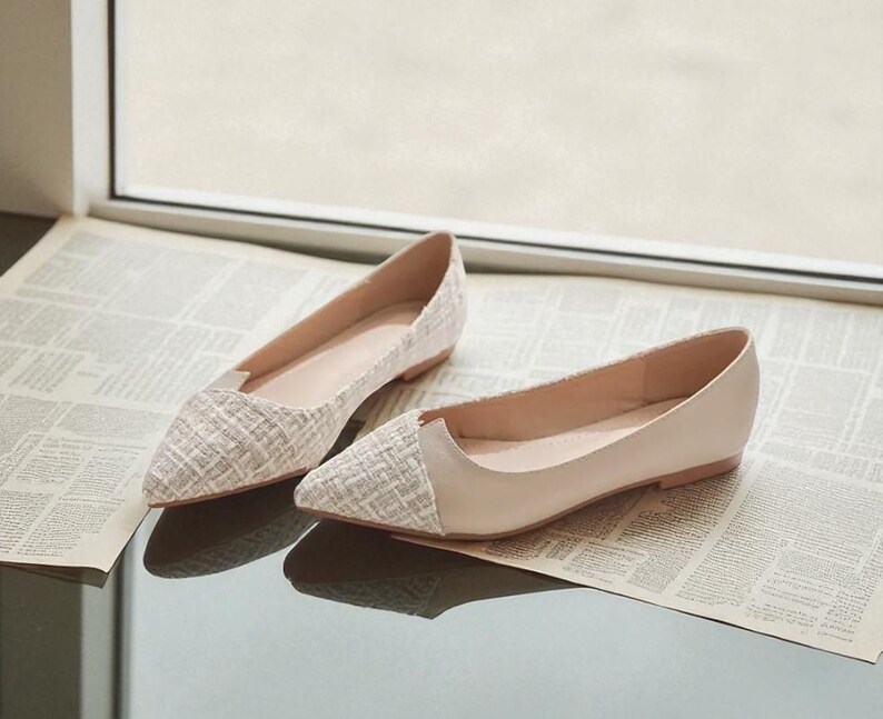 May include: A pair of beige and white flats with a pointed toe. The toe of each shoe has a textured, woven pattern, while the rest of the shoe is a smooth, solid beige. The shoes are displayed on a newspaper.