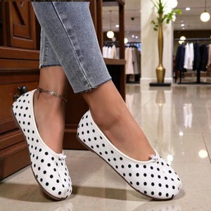 Könnte beinhalten: Weiße Ballerinas mit schwarzen Polka Dots. Die Schuhe haben eine runde Spitze und eine kleine Schleife oben. Die Sohlen sind braun. Die Schuhe werden mit einer blauen Jeans getragen.