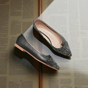 May include: A pair of black flats with a pointed toe and a woven, textured toe cap. The shoes have a low heel and a light brown sole. The shoes are displayed on a vintage newspaper background.