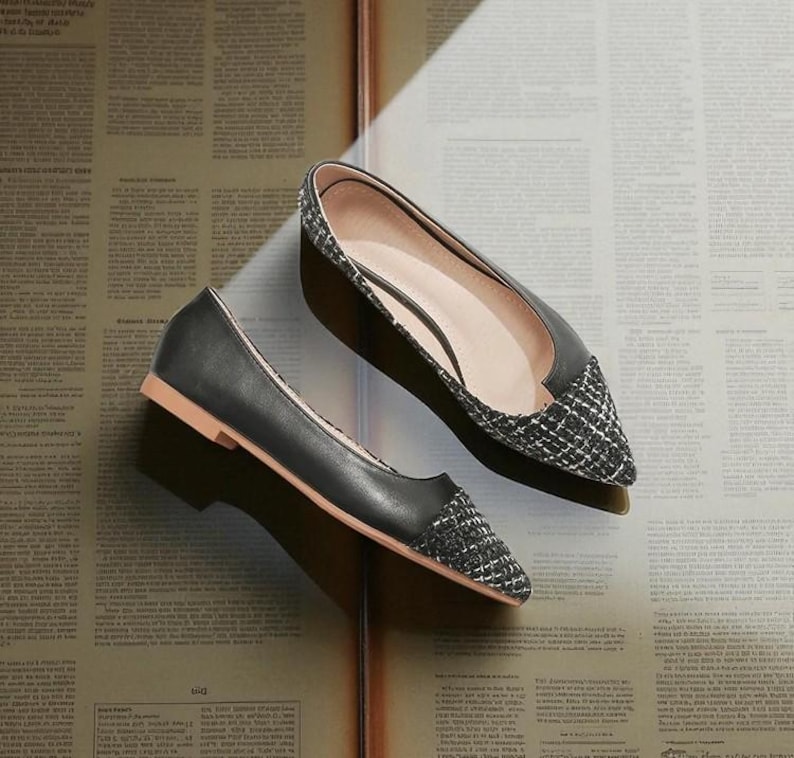 May include: A pair of black flats with a pointed toe and a woven, textured toe cap. The shoes have a low heel and a light brown sole. The shoes are displayed on a vintage newspaper background.