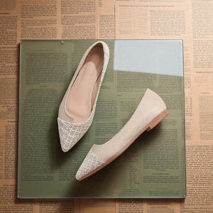 May include: A pair of beige flats with a pointed toe and a woven texture on the toe cap. The shoes are displayed on a glass surface with a green tint, set against a backdrop of vintage newspaper clippings.