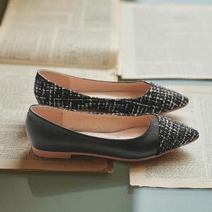 May include: A pair of black and white tweed and black leather flats. The shoes have a pointed toe design and are set on a light brown sole. The shoes are displayed on top of open newspapers.