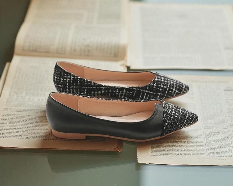 May include: A pair of black and white tweed and black leather flats. The shoes have a pointed toe design and are set on a light brown sole. The shoes are displayed on top of open newspapers.