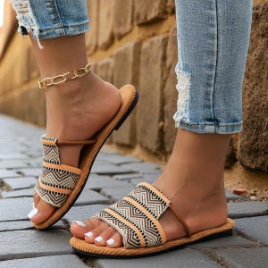 May include: Sandals with a geometric pattern in beige, black, and brown. The open-toe slides have a flat sole and a strap that goes between the toes. The sandals are worn with blue jeans and a gold chain ankle bracelet.