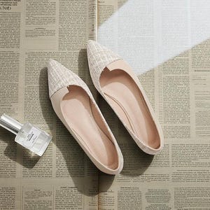 May include: A pair of beige flats with a pointed toe and a textured fabric detail on the toe cap. The shoes are displayed on a newspaper background, next to a perfume bottle with the word "COLOGNE" visible.