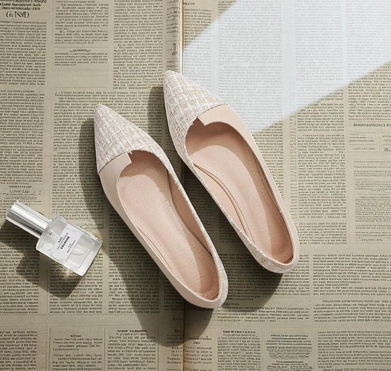 May include: A pair of beige flats with a pointed toe and a textured fabric detail on the toe cap. The shoes are displayed on a newspaper background, next to a perfume bottle with the word "COLOGNE" visible.