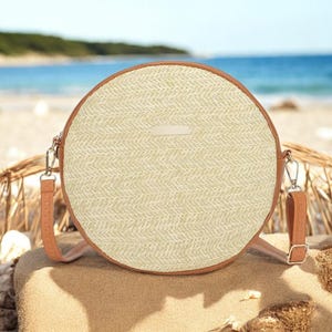 Pode incluir: Uma bolsa redonda de palha com acabamento em couro marrom e uma alça longa de couro marrom. A bolsa está sentada em uma praia de areia.