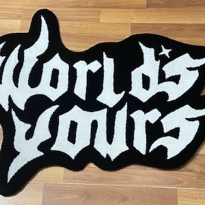 May include: Black and white rug with the words "Worlds Yours" in a gothic font. The rug has a unique, irregular shape and a star design. The rug is placed on a wooden floor.