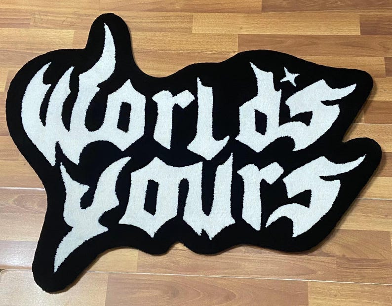 May include: Black and white rug with the words "Worlds Yours" in a gothic font. The rug has a unique, irregular shape and a star design. The rug is placed on a wooden floor.