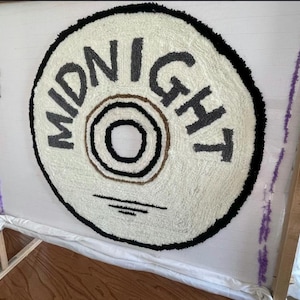 May include: A handmade rug in progress, featuring a circular design with the word "MIDNIGHT" in gray letters. The rug has a cream-colored background with a black border and a brown and black center. The rug is mounted on a wooden frame.