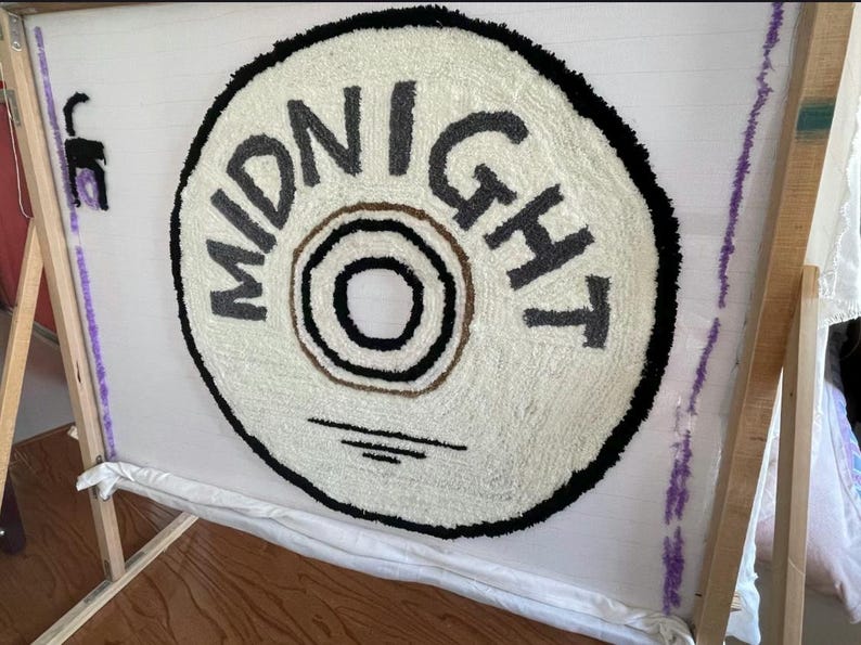May include: A handmade rug in progress, featuring a circular design with the word "MIDNIGHT" in gray letters. The rug has a cream-colored background with a black border and a brown and black center. The rug is mounted on a wooden frame.