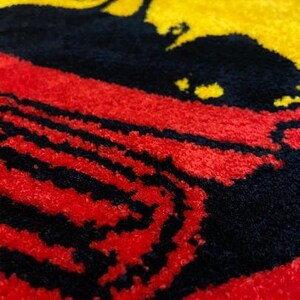 May include: A close-up of a textured, colorful rug with a silhouette of a person on a yellow background. The rug features a red and black striped pattern, creating a bold and eye-catching design. The rug is likely made of a soft material.