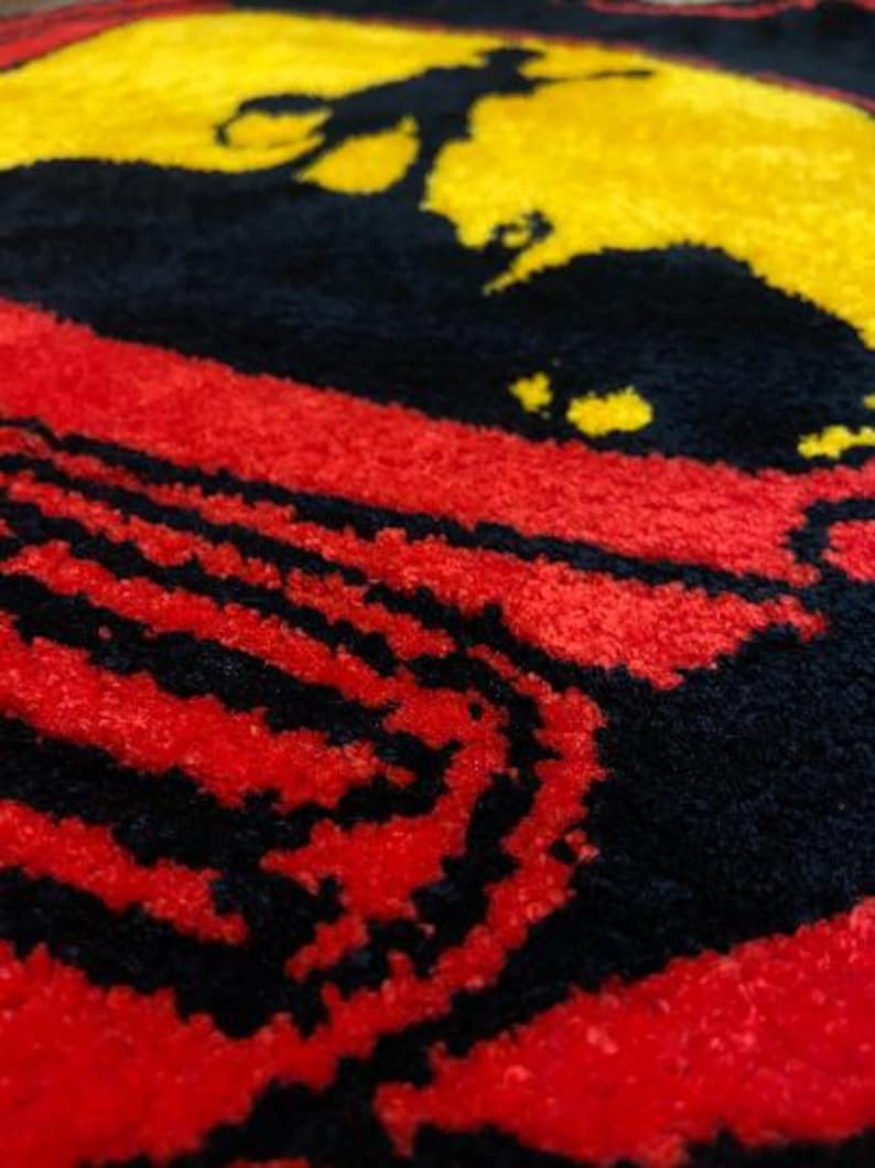 May include: A close-up of a textured, colorful rug with a silhouette of a person on a yellow background. The rug features a red and black striped pattern, creating a bold and eye-catching design. The rug is likely made of a soft material.